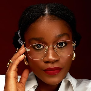 May include: A woman wearing gold-rimmed, rimless eyeglasses and gold hoop earrings. She is also wearing gold rings. Her lips are painted red, and she is wearing a white collared shirt. The background is a dark red.