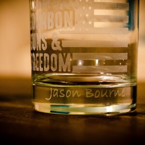 May include: A clear glass tumbler with an etched design of an American flag and the words "Bourbon, Guns & Freedom". The bottom of the glass is etched with the name "Jason Bourne".