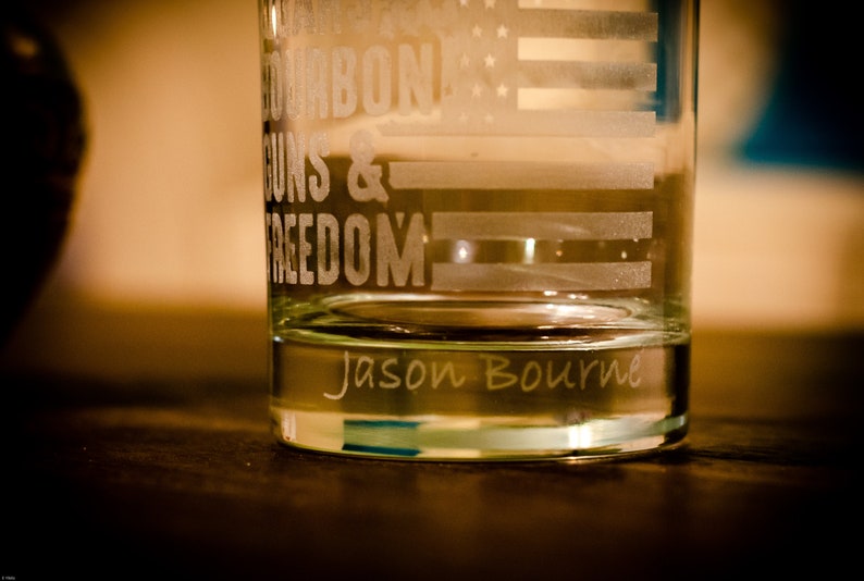 May include: A clear glass tumbler with an etched design of an American flag and the words "Bourbon, Guns & Freedom". The bottom of the glass is etched with the name "Jason Bourne".