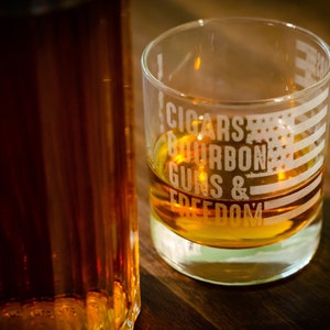 May include: A clear glass with an American flag design and the text "CIGARS BOURBON GUNS & FREEDOM" etched on the side. The glass is filled with a golden brown liquid.