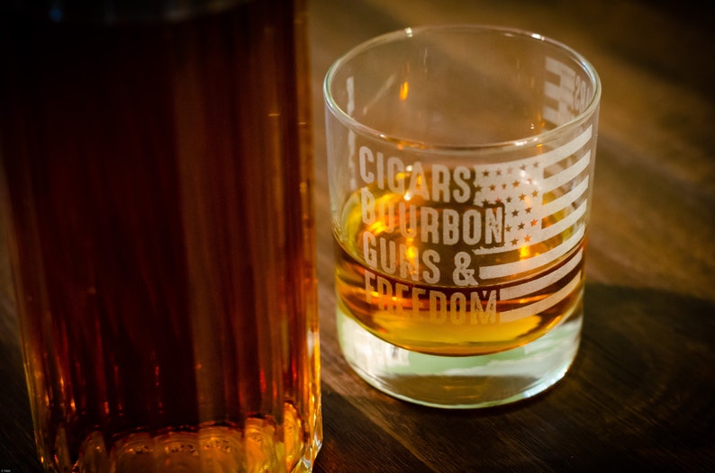May include: A clear glass with an American flag design and the text "CIGARS BOURBON GUNS & FREEDOM" etched on the side. The glass is filled with a golden brown liquid.