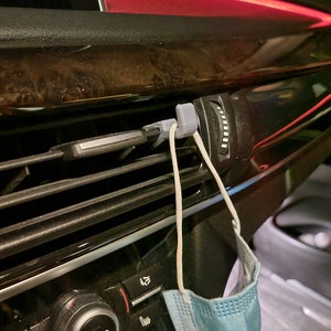 May include: A gray 3D-printed car vent clip holds a light blue face mask. The clip is attached to the car's air vent, with a red light strip visible in the background. The car's interior features black and brown trim.