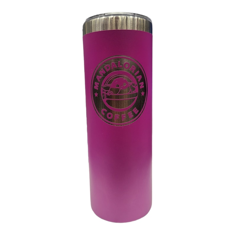 May include: A tall, pink tumbler with a silver lid. The tumbler features a gold-colored emblem with the words "MANDALORIAN COFFEE" and an image of Grogu. The tumbler is designed to keep beverages hot or cold.