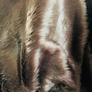 May include: Close-up of a brown faux fur fabric. The fabric has a soft, textured appearance with long, flowing fibers. The color varies from light to dark brown, creating a natural, shaded effect. The material appears to be plush and luxurious.