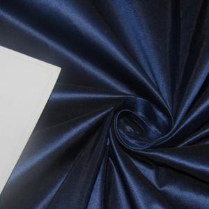 May include: A close-up of a navy blue fabric with a satin sheen. The fabric is draped and swirled, creating folds and shadows that highlight its texture. A white rectangular shape is in the upper left corner.