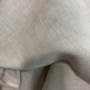 May include: Close-up of a beige linen fabric. The fabric has a subtle texture and a natural, slightly wrinkled appearance. The folds and shadows create depth, highlighting the weave of the material. The color is a neutral, warm tone.