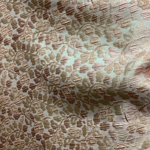 May include: Close-up of a fabric with a repeating floral pattern. The fabric features a textured surface with a mix of colours, including light pink, beige, and gold. The design has a subtle sheen, adding to its visual appeal.