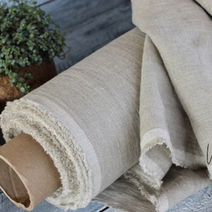 May include: A roll of natural linen fabric, showcasing a light beige color with a subtle texture. The fabric is rolled around a cardboard tube, with a frayed edge detail. A small potted plant is visible in the background.