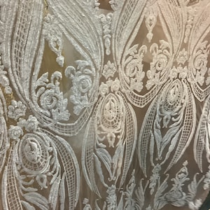 Puede incluir: Tela blanca bordada con un patrón floral sobre un fondo nude. La tela es transparente y tiene un diseño delicado e intrincado.
