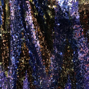 May include: Close-up of iridescent blue, gold, and purple sequins arranged in a wavy pattern. The sequins are sewn onto a fabric base.