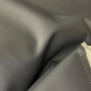 May include: Close-up of a dark gray, possibly black, fabric with soft folds and a smooth texture. The fabric appears to be a solid color, with subtle variations in tone due to the light. The image suggests a textile or material.