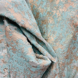 以下が含まれることがあります： テクスチャのある抽象的なパターンが施された、ターコイズとゴールドのファブリックのクローズアップ。ファブリックは折り畳まれ、ドレープされており、デザインの複雑なディテールがわかります。