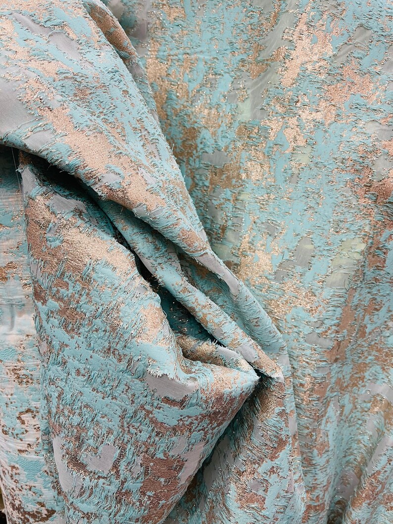 以下が含まれることがあります： テクスチャのある抽象的なパターンが施された、ターコイズとゴールドのファブリックのクローズアップ。ファブリックは折り畳まれ、ドレープされており、デザインの複雑なディテールがわかります。