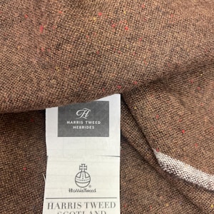 May include: Close-up of brown Harris Tweed fabric with flecks of red and yellow. A tag reads "Harris Tweed Hebrides" and "Harris Tweed Scotland". The fabric is tightly woven and appears to be a wool blend.