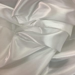 Può includere: Primo piano di tessuto di raso bianco. Il tessuto è drappeggiato e piegato, creando ombre morbide e riflessi. Il materiale ha una consistenza liscia e lucida, che riflette la luce. L'effetto complessivo è elegante e fluido.