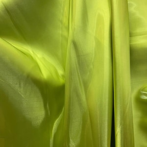 May include: Close-up of a vibrant, lime green fabric with a silky, shiny texture. The fabric is draped, creating soft folds and shadows. The material appears lightweight and flowing, suggesting it could be used for clothing or decorative purposes.