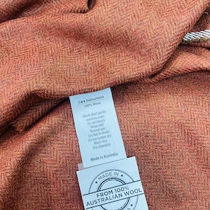 Puede incluir: Una etiqueta blanca con texto negro unida a una tela marrón y naranja. La etiqueta dice "Care Instructions 100% Wool" y enumera las instrucciones de lavado. Una segunda etiqueta blanca con texto negro dice "Made in * FROM 100% AUSTRALIAN WOOL * AUSTRALIA *"