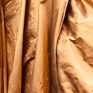 May include: Close-up of a draped fabric in a warm, golden-brown hue. The material has a subtle sheen and visible folds, creating a textured appearance. The light catches the fabric, highlighting its rich color and creating shadows.