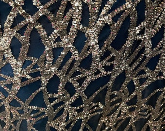 May include: Close-up of a navy blue fabric with a silver sequin pattern. The sequins are arranged in a geometric design, creating a shimmering effect.
