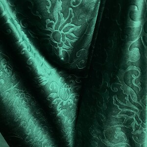 May include: Dark green fabric with a floral pattern. The fabric has a velvety texture and is draped in soft folds, creating shadows and highlights. The floral design is embossed, adding depth and dimension to the material.