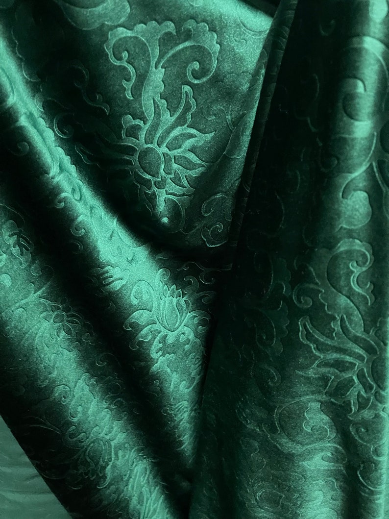 May include: Dark green fabric with a floral pattern. The fabric has a velvety texture and is draped in soft folds, creating shadows and highlights. The floral design is embossed, adding depth and dimension to the material.