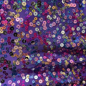 May include: Close-up of a purple fabric covered in iridescent sequins. The sequins are round and vary in size, reflecting a rainbow of colors including pink, gold, and blue. The fabric is slightly gathered, creating texture and dimension.