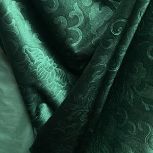 May include: Close-up of a dark teal fabric with an embossed floral pattern. The fabric is folded, creating shadows and highlights that emphasize the texture and design. The overall effect is luxurious and elegant.