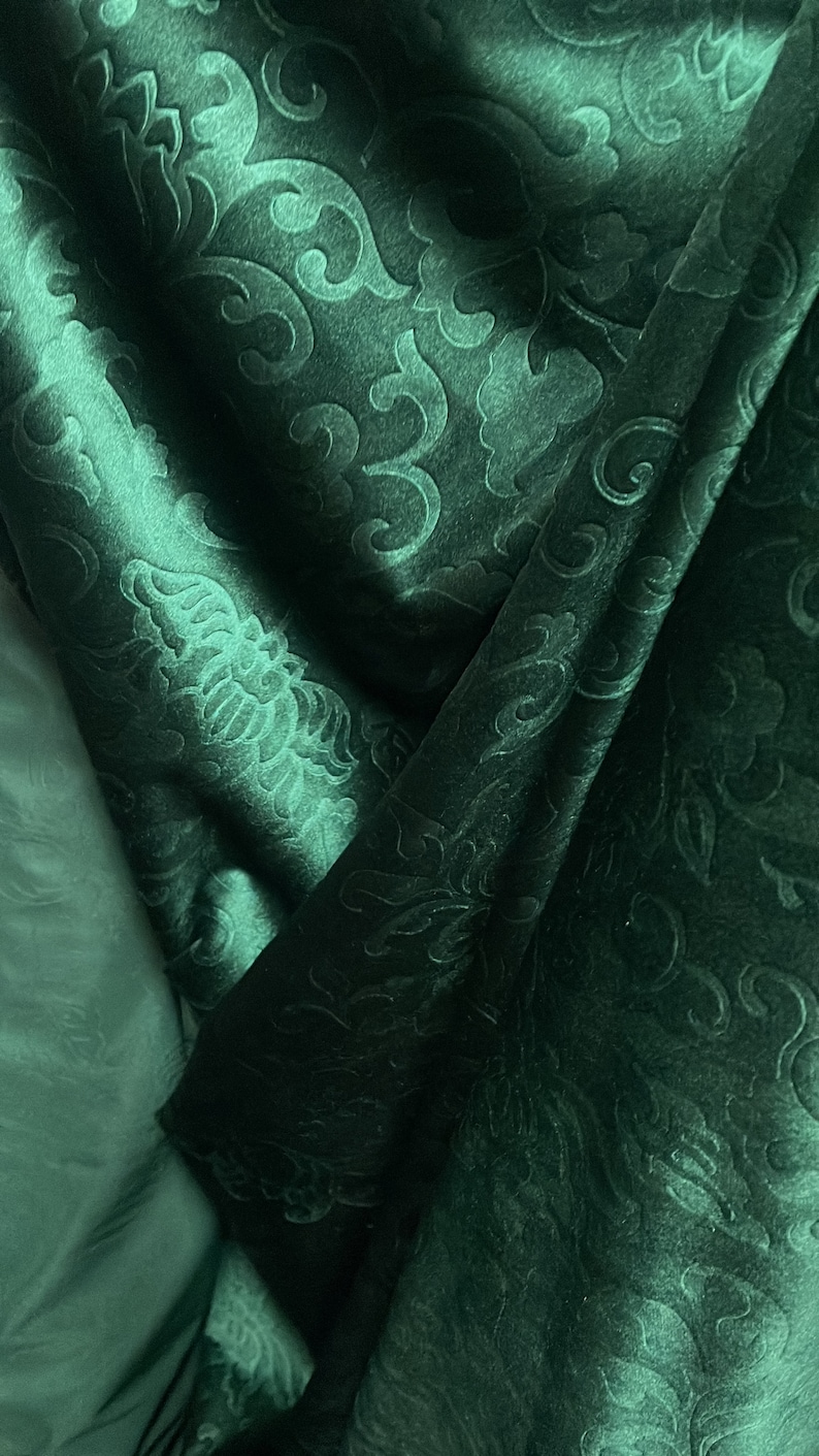 May include: Close-up of a dark teal fabric with an embossed floral pattern. The fabric is folded, creating shadows and highlights that emphasize the texture and design. The overall effect is luxurious and elegant.