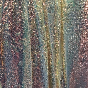 May include: Close-up of iridescent fabric with a textured, scaly pattern. The fabric is made of sequins and has a light blue, white, and gold color scheme.
