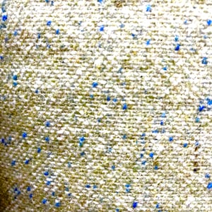 May include: Close-up of a textured fabric with a speckled pattern. The fabric has a light beige base with flecks of green and small, scattered blue dots. The weave appears tight and durable, suggesting a sturdy material.