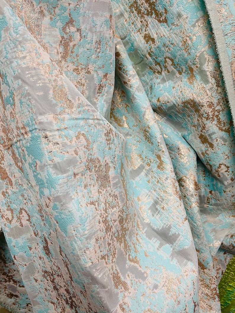 以下が含まれることがあります： テクスチャのある抽象的なパターンが施された、ティールとゴールドのファブリックのクローズアップ。ファブリックはメタリックな光沢があり、柔らかくドレープ感のある感触です。