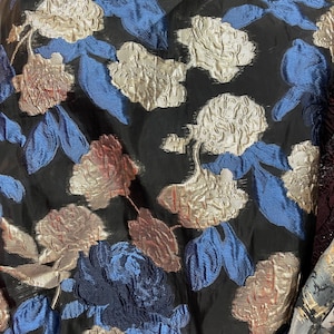 May include: Close-up of a black fabric with a floral pattern. The design features gold, blue, and rust-colored flowers and leaves. The fabric has a textured appearance, with a slight sheen.