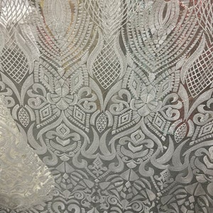 Könnte beinhalten: Nahaufnahme eines weißen Spitzenstoffs mit einem komplizierten, sich wiederholenden Muster. Das Design zeigt florale und geometrische Formen, darunter Diamanten, Wirbel und stilisierte Blätter. Der Stoff wirkt zart und halbtransparent.