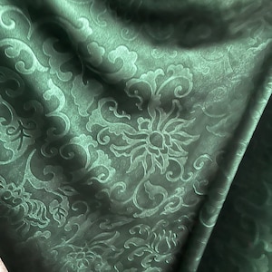 May include: Dark green fabric with a floral pattern. The fabric has a subtle sheen and a raised, embossed design featuring stylized flowers and swirling leaves. The folds in the fabric create shadows that highlight the texture and depth of the pattern.