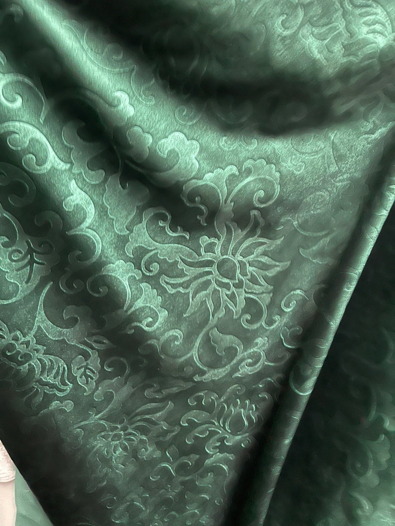 May include: Dark green fabric with a floral pattern. The fabric has a subtle sheen and a raised, embossed design featuring stylized flowers and swirling leaves. The folds in the fabric create shadows that highlight the texture and depth of the pattern.