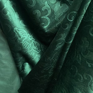 May include: Close-up of a dark teal fabric with an embossed floral pattern. The fabric is folded, creating shadows and highlighting the texture. The design features stylized flowers and swirling leaves.