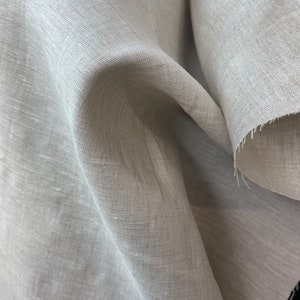 May include: Close-up of a light gray linen fabric. The fabric is loosely woven, with visible texture and a slightly wrinkled appearance. The edges of the fabric are unfinished, showing the raw, frayed threads. The overall look is natural and soft.