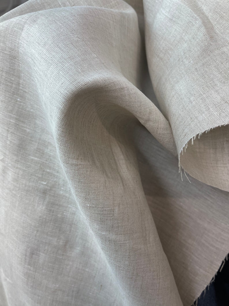 May include: Close-up of a light gray linen fabric. The fabric is loosely woven, with visible texture and a slightly wrinkled appearance. The edges of the fabric are unfinished, showing the raw, frayed threads. The overall look is natural and soft.