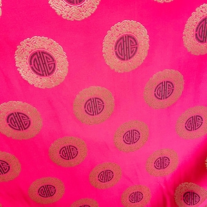 May include: A pink fabric with a repeating pattern of gold and black circles. Each circle contains a stylized Chinese character.
