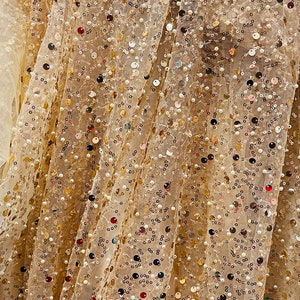 May include: Close-up of a beige fabric with gold, silver, and iridescent sequins and small white beads. The fabric is draped and folded, creating a textured and sparkly surface.