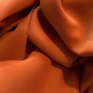 May include: Close-up of a rust-colored fabric with a smooth, silky texture. The fabric is folded and draped, creating soft, flowing lines.