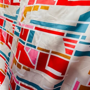 Könnte beinhalten: Ein weißer Stoff mit einem geometrischen Muster in Rot, Rosa, Blau und Gold. Der Stoff ist gefaltet und hat eine strukturierte Oberfläche.