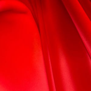 May include: A close-up shot of vibrant red fabric with soft folds and subtle shadows. The material appears smooth and luxurious, with a slight sheen, suggesting a satin or silk texture. The image highlights the rich color and flowing drape of the fabric.
