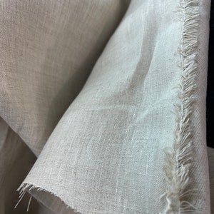 May include: Close-up of a beige linen fabric with a raw, fringed edge. The fabric is tightly woven, with visible texture and subtle variations in color. The folds and shadows create depth, highlighting the natural drape of the material.