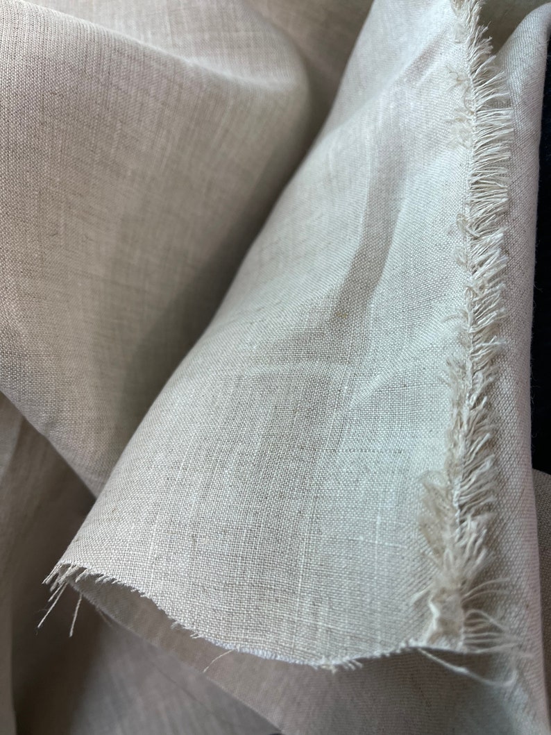 May include: Close-up of a beige linen fabric with a raw, fringed edge. The fabric is tightly woven, with visible texture and subtle variations in color. The folds and shadows create depth, highlighting the natural drape of the material.