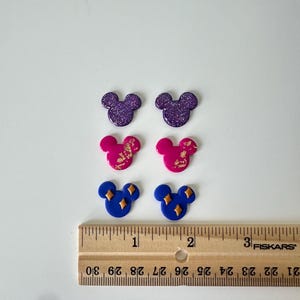 May include: Six colourful Mickey Mouse-shaped earrings in purple glitter, pink with gold flecks, and blue with gold diamond accents. The earrings are arranged on a white surface next to a ruler, measuring approximately 2.5 cm in length.