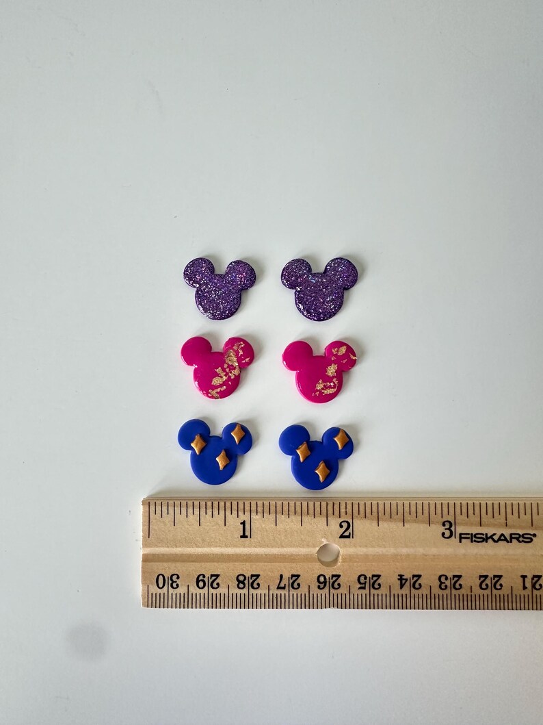 May include: Six colourful Mickey Mouse-shaped earrings in purple glitter, pink with gold flecks, and blue with gold diamond accents. The earrings are arranged on a white surface next to a ruler, measuring approximately 2.5 cm in length.