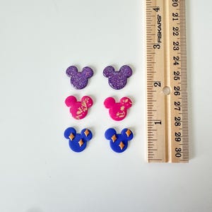 May include: Six Mickey Mouse-shaped earrings in various colours. Two are purple with glitter, two are pink with gold accents, and two are blue with gold diamond shapes. A wooden ruler is in the background.