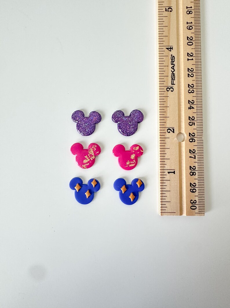 May include: Six Mickey Mouse-shaped earrings in various colours. Two are purple with glitter, two are pink with gold accents, and two are blue with gold diamond shapes. A wooden ruler is in the background.