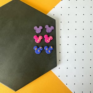 May include: Six colourful Mickey Mouse-shaped stud earrings. Two are purple with glitter, two are pink with gold accents, and two are blue with yellow diamond accents. The earrings are arranged on a dark grey hexagonal surface against a yellow and white background.